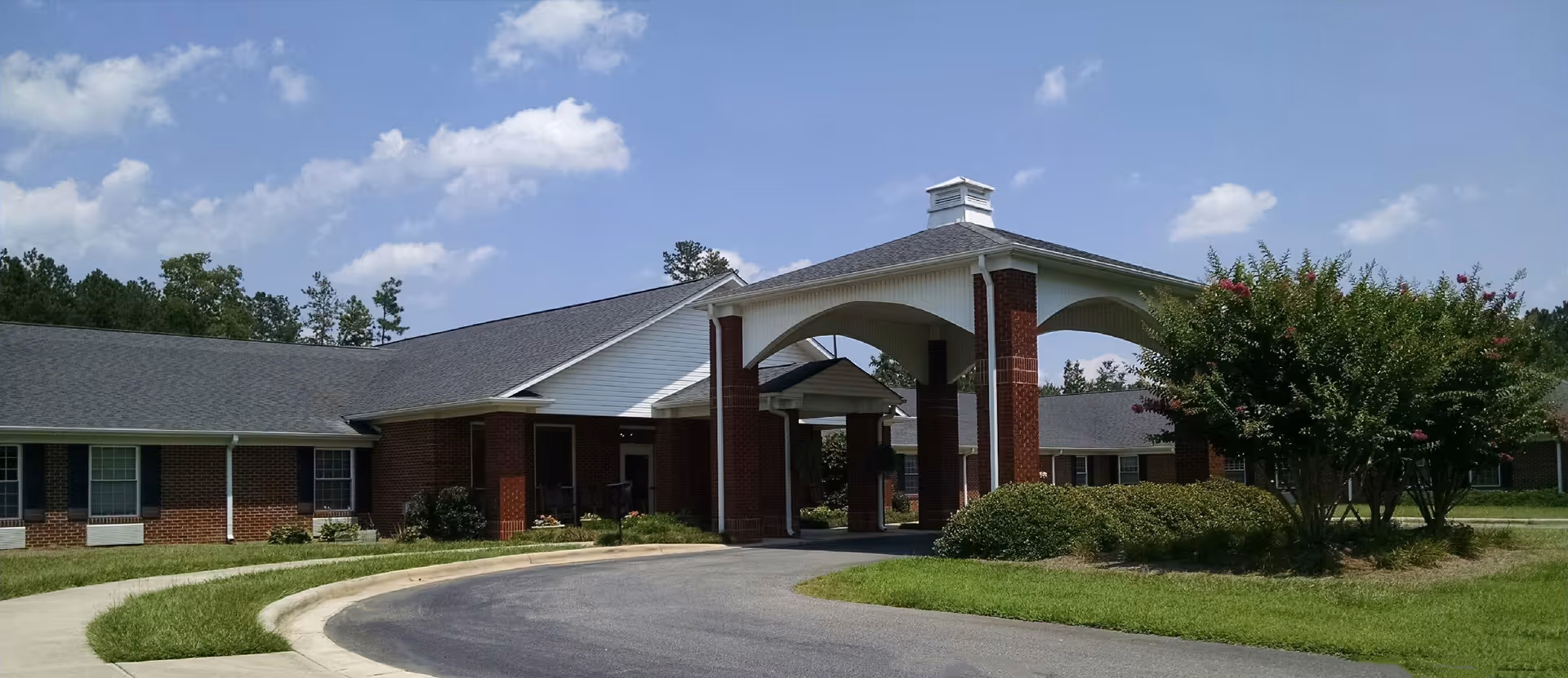 Front exterior of a single-story brick senior living facility with a covered porte-cochere, curved driveway, and landscaped lawn.
