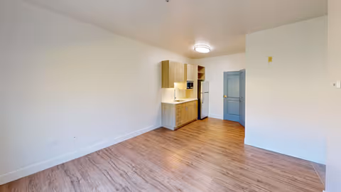 Empty apartment room with wood-look flooring and a small kitchenette with cabinets, sink and refrigerator along the back wall.