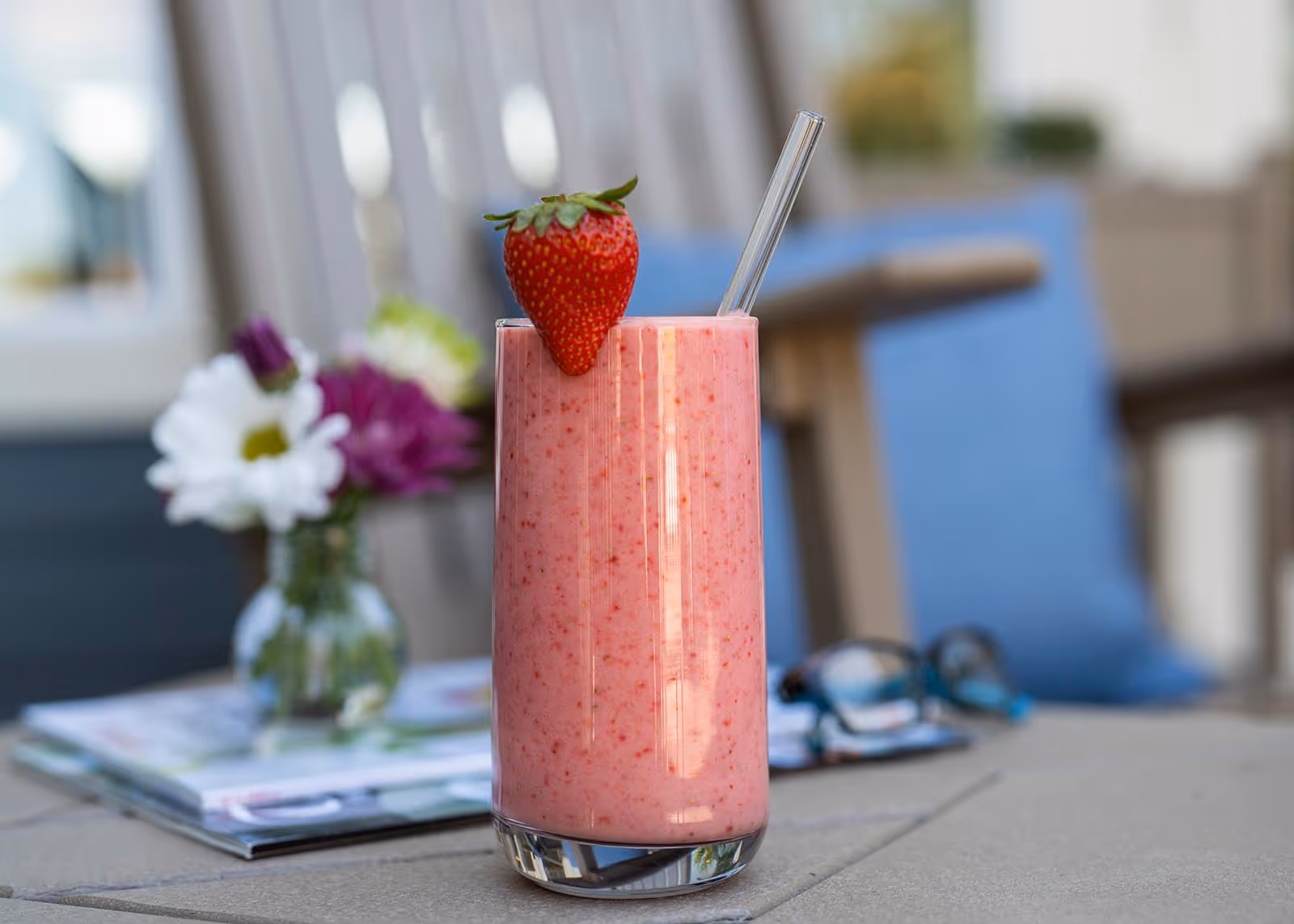A tall glass of pink strawberry smoothie with a fresh strawberry garnish and a clear straw, placed on a table. In the background, there is a small vase with white and purple flowers, a pair of eyeglasses, and some magazines, with blurred outdoor seating visible.