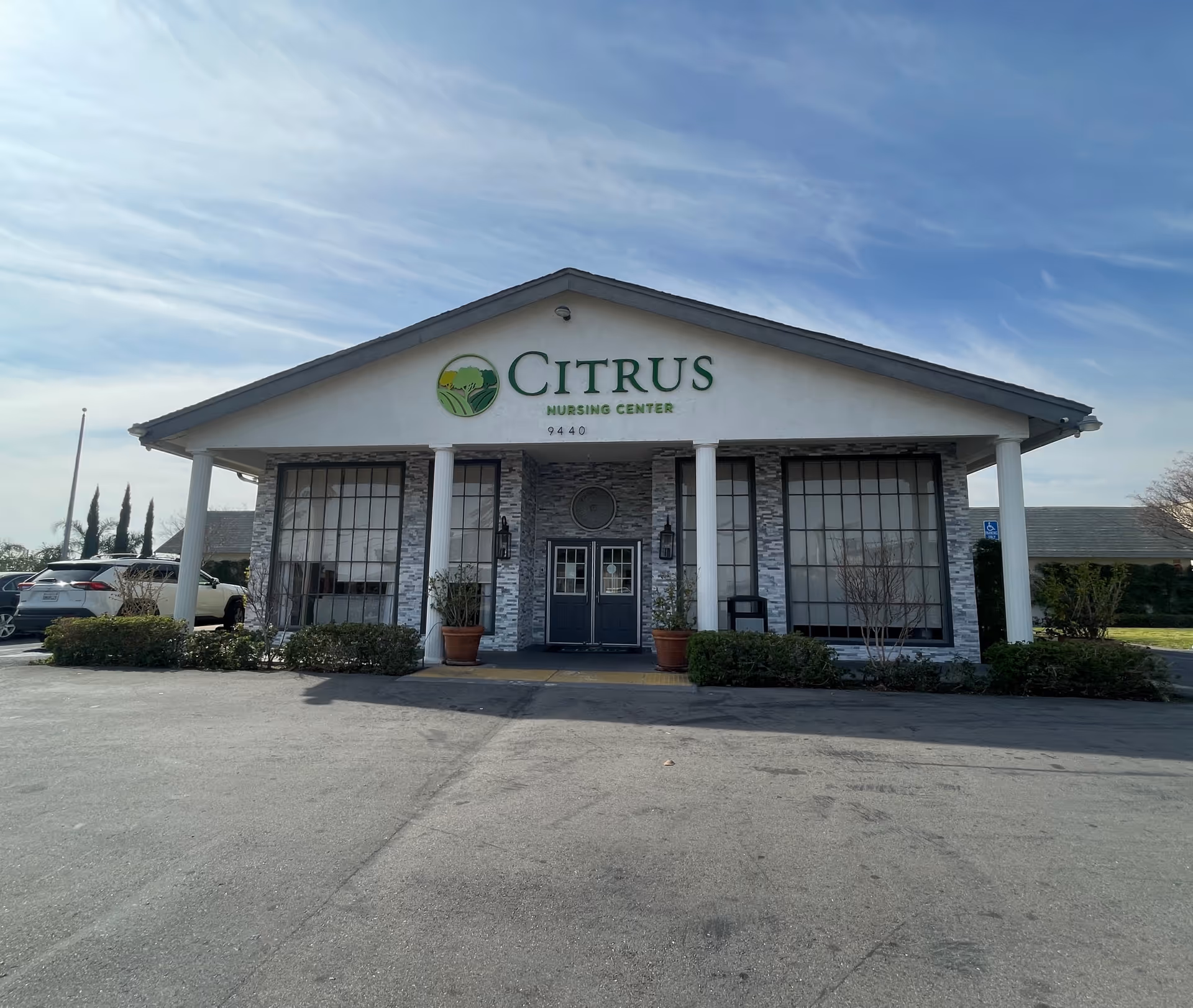 Front exterior view of a single-story building with a peaked roof and large windows. The building has a sign that reads 'Citrus Nursing Center' above the entrance, which features double doors flanked by two white columns and potted plants. There are cars parked to the left and some greenery around the building under a partly cloudy sky.