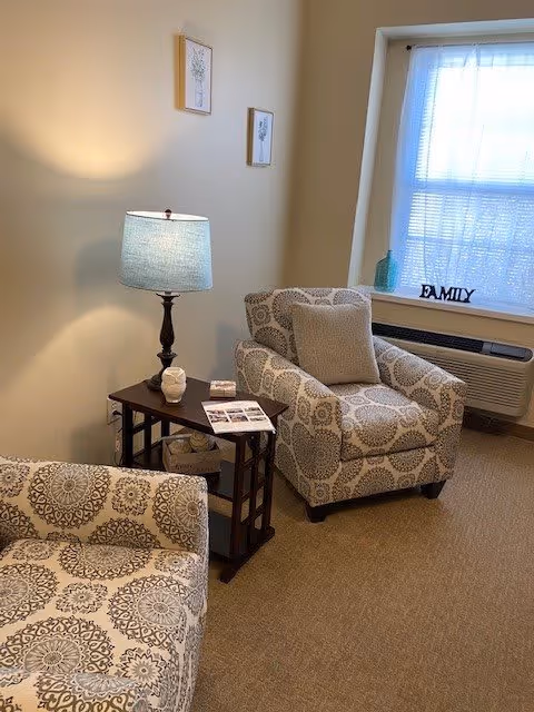 A cozy living room area with two patterned armchairs and a matching sofa. A wooden side table with a lamp, a small decorative item, and some reading material sits between the chairs. A window with sheer white curtains lets in natural light, and a decorative 'FAMILY' sign is placed on the windowsill above an air conditioning unit.