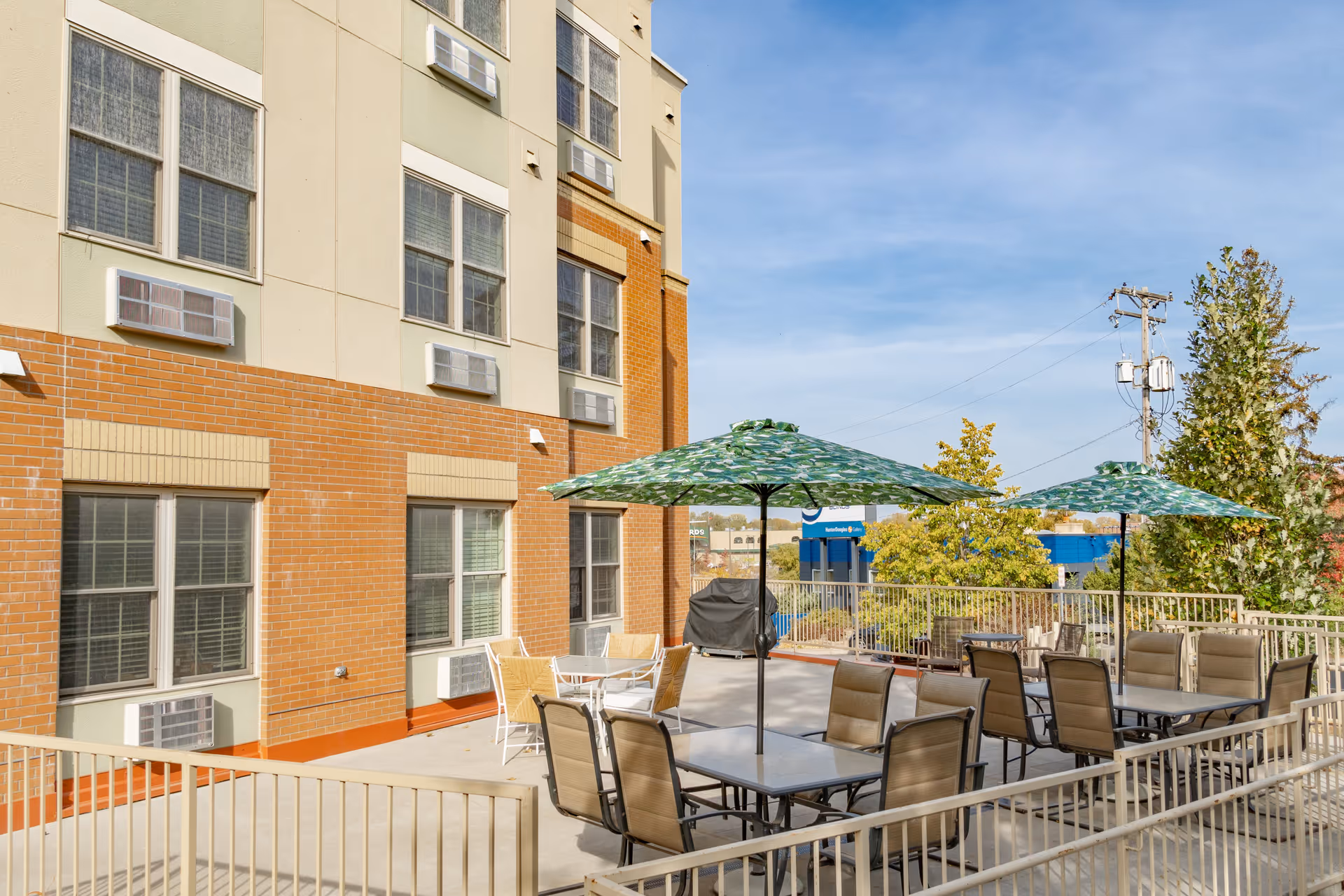 Outdoor patio area at Cedar Ridge Place with multiple tables and chairs under green patterned umbrellas, adjacent to a multi-story building with windows and air conditioning units. Trees and a blue building are visible in the background under a clear sky.