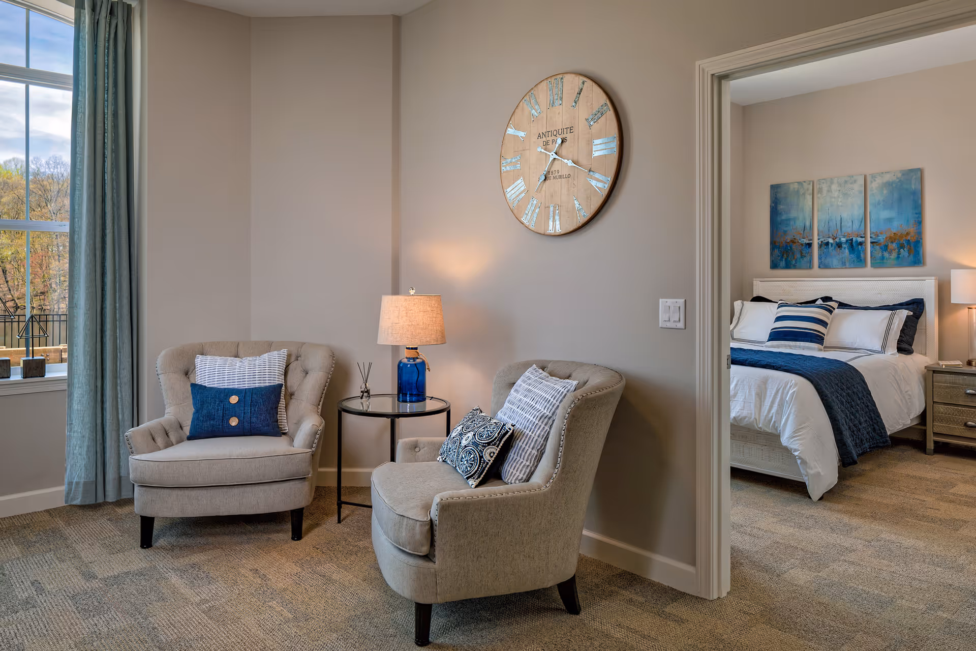 A cozy sitting area with two beige armchairs, each adorned with decorative pillows, and a small round side table with a blue glass lamp. A large round wall clock hangs above the chairs. Through an open doorway, a bedroom is visible with a neatly made bed featuring white and navy blue bedding, a nightstand with a lamp, and a three-panel blue artwork above the headboard.
