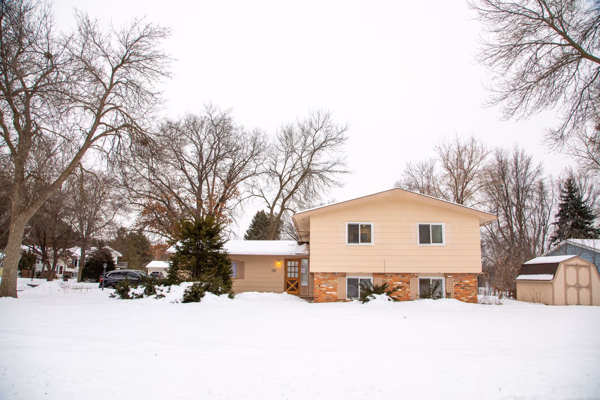 Snow-covered front yard with a split-level house, bare trees, and a small shed.