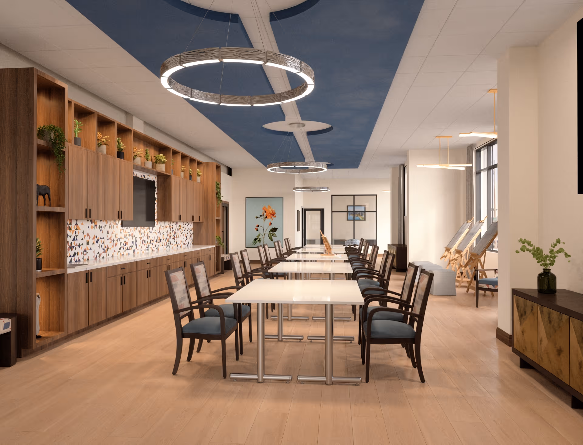A spacious, well-lit dining room with multiple tables and chairs arranged in rows. The room features wooden cabinetry along one wall with decorative plants and a colorful tiled backsplash. Modern circular pendant lights hang from the ceiling, which has a blue accent design. Large windows on the right side let in natural light, and there are easels and seating near the windows. A sideboard with a vase of greenery is visible on the right.