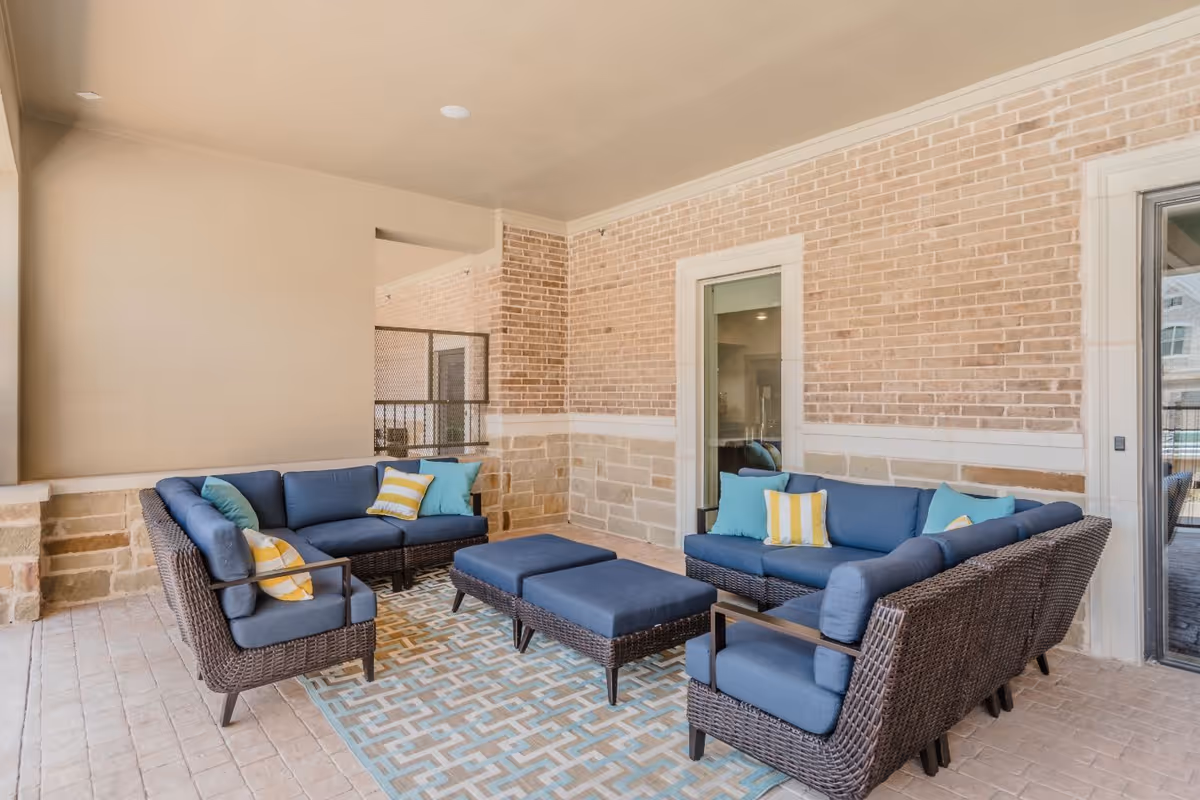 Covered outdoor patio with wicker sectional sofas and blue cushions arranged around a central ottoman on a patterned rug against a brick wall.