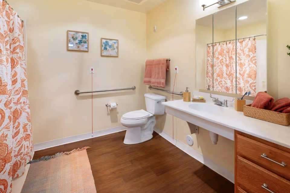 A clean and accessible bathroom with a white toilet, a long white countertop with a sink, a large mirror above the sink, and a shower with a floral-patterned curtain. The bathroom has wooden flooring, beige walls, two framed floral pictures on the wall, a towel rack with a pink towel, and a basket with folded towels on the countertop.