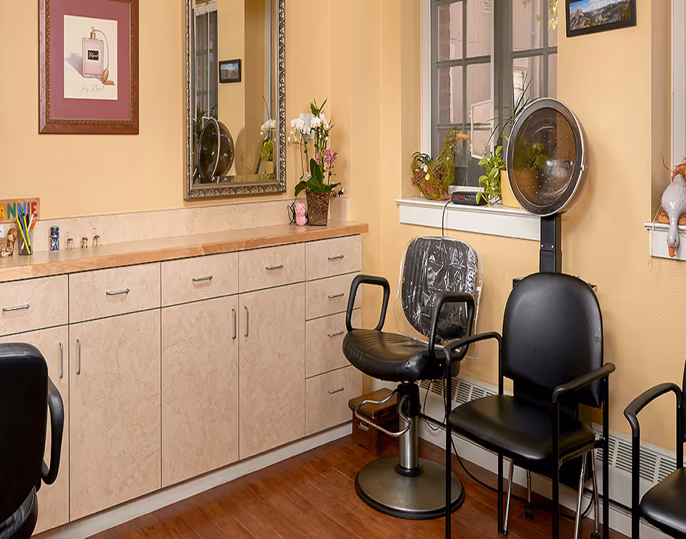 Interior view of a salon area in a senior living facility with beige walls and wooden flooring. There is a long counter with multiple drawers and cabinets, a large decorative mirror above it, and a framed picture on the wall. Two black salon chairs are positioned near a window, one with a plastic cover and a hair dryer hood attached behind it. Various decorative plants and items are placed on the counter and windowsill.