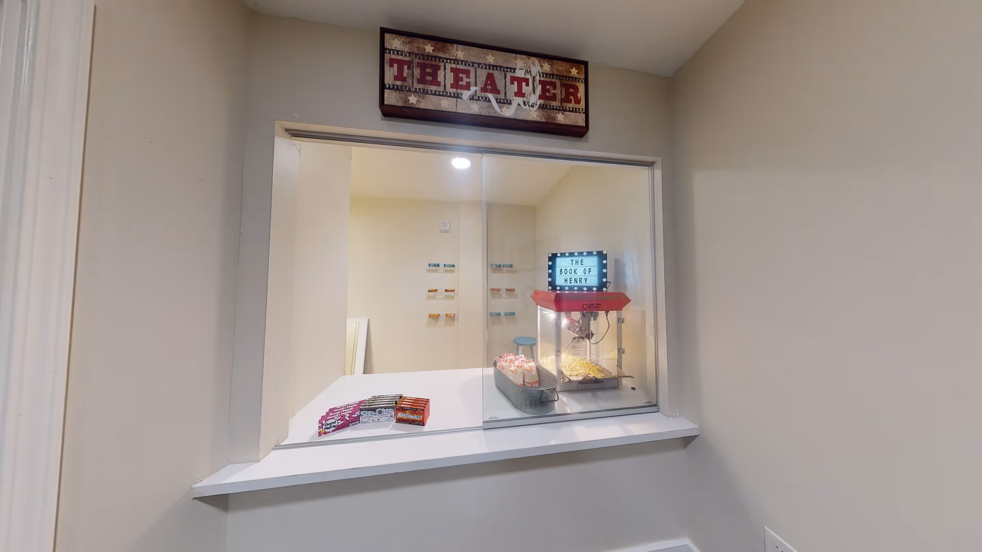A small theater concession window with a popcorn machine, popcorn bags, and candy boxes. Above the window is a sign that reads 'THEATER'. Inside the concession area, a lightbox sign displays the text 'THE BOOK OF HENRY'.