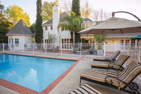 Outdoor swimming pool area with striped lounge chairs and a large umbrella beside the pool. The pool is surrounded by a white safety fence, and in the background, there is a large white building with multiple windows and tall trees.