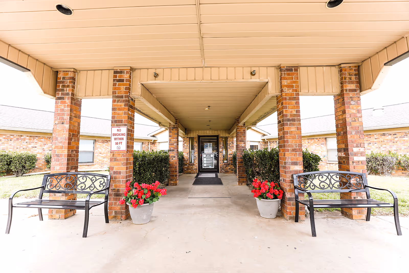 Covered entrance with brick columns, benches, and potted red flowers leading to double glass doors of a senior living facility.