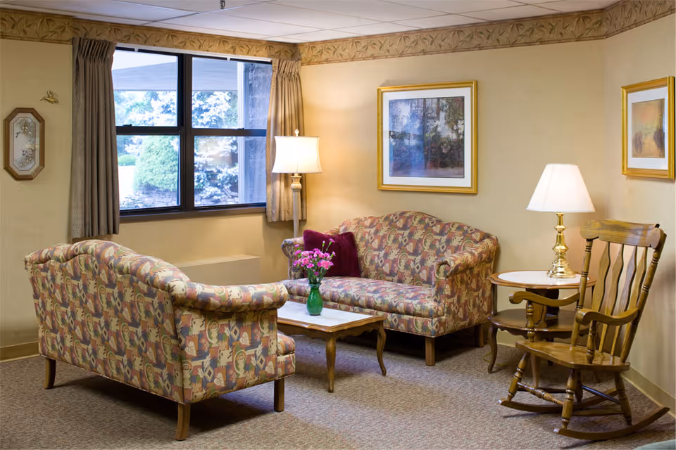 A cozy living room area with two patterned sofas facing each other, a wooden coffee table with a green vase of pink flowers, a wooden rocking chair, two side tables with lamps, framed artwork on beige walls, and a window with curtains showing greenery outside.