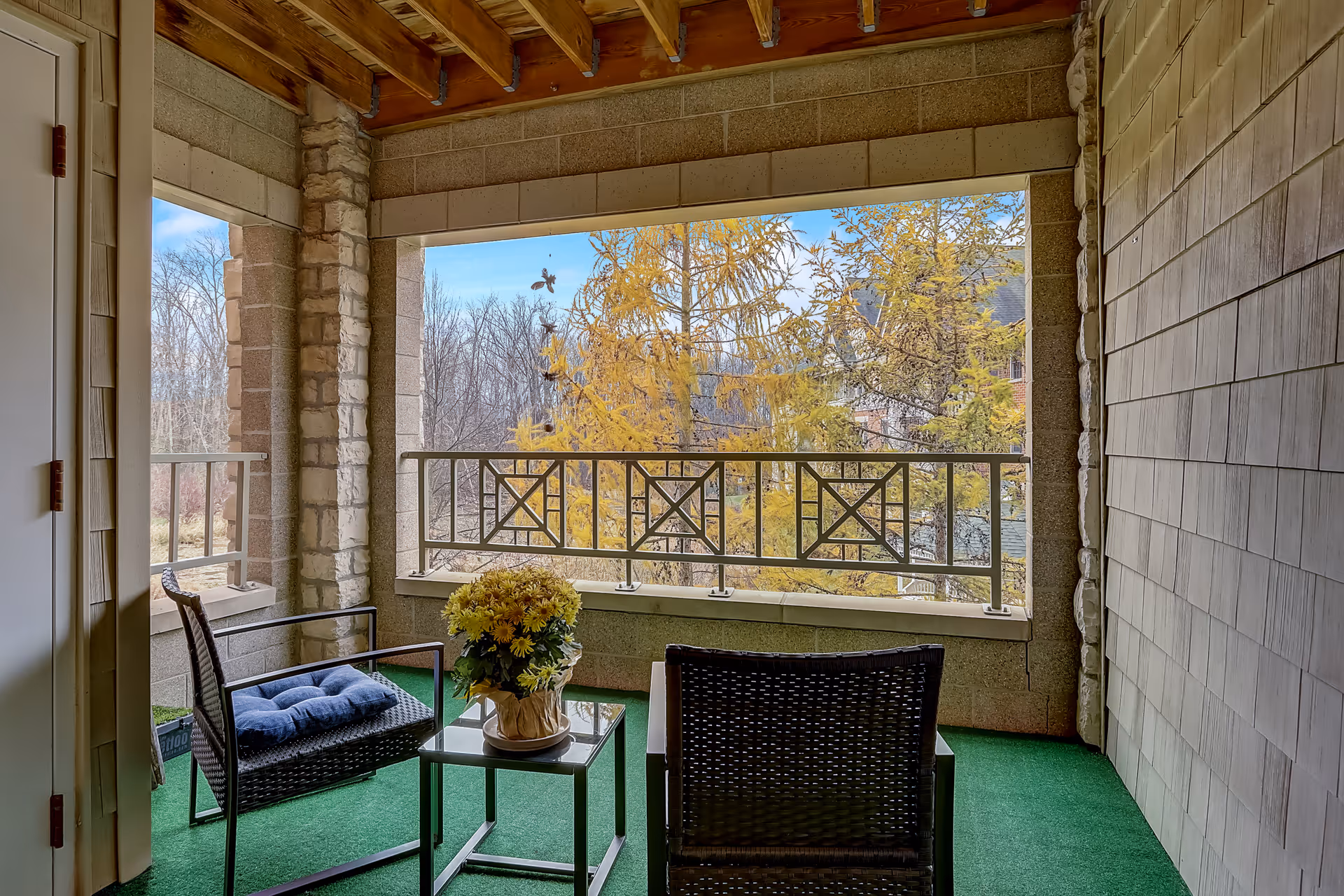 Covered balcony area with two black wicker chairs, one with a blue cushion, and a small glass table with a potted yellow flower arrangement. The balcony has a green carpet floor, stone and wood ceiling, and a metal railing with a view of trees with yellow leaves and a clear blue sky.