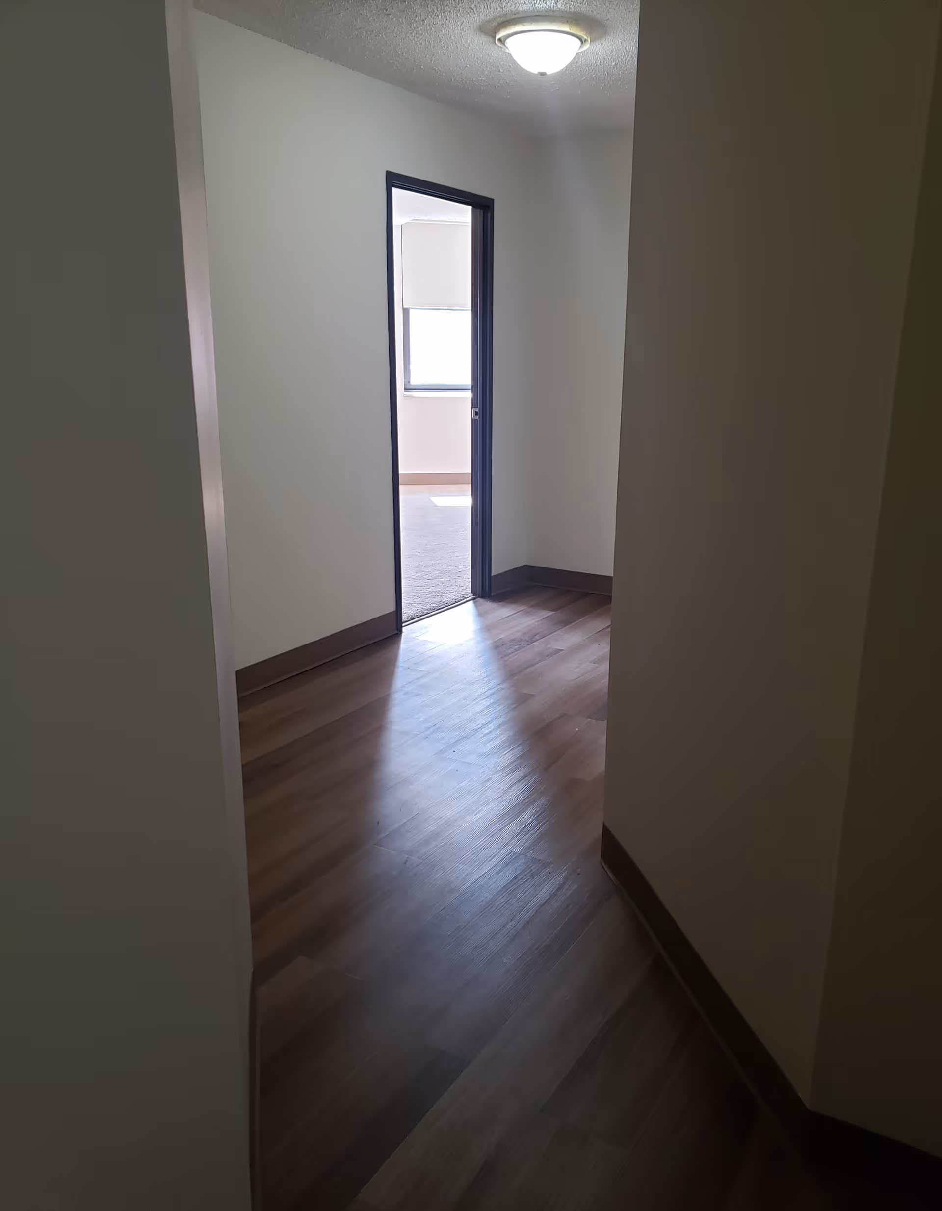 A hallway with wooden flooring leading to a room with a window. The walls are painted white with dark brown baseboards and door trim. A ceiling light fixture is visible above the hallway.