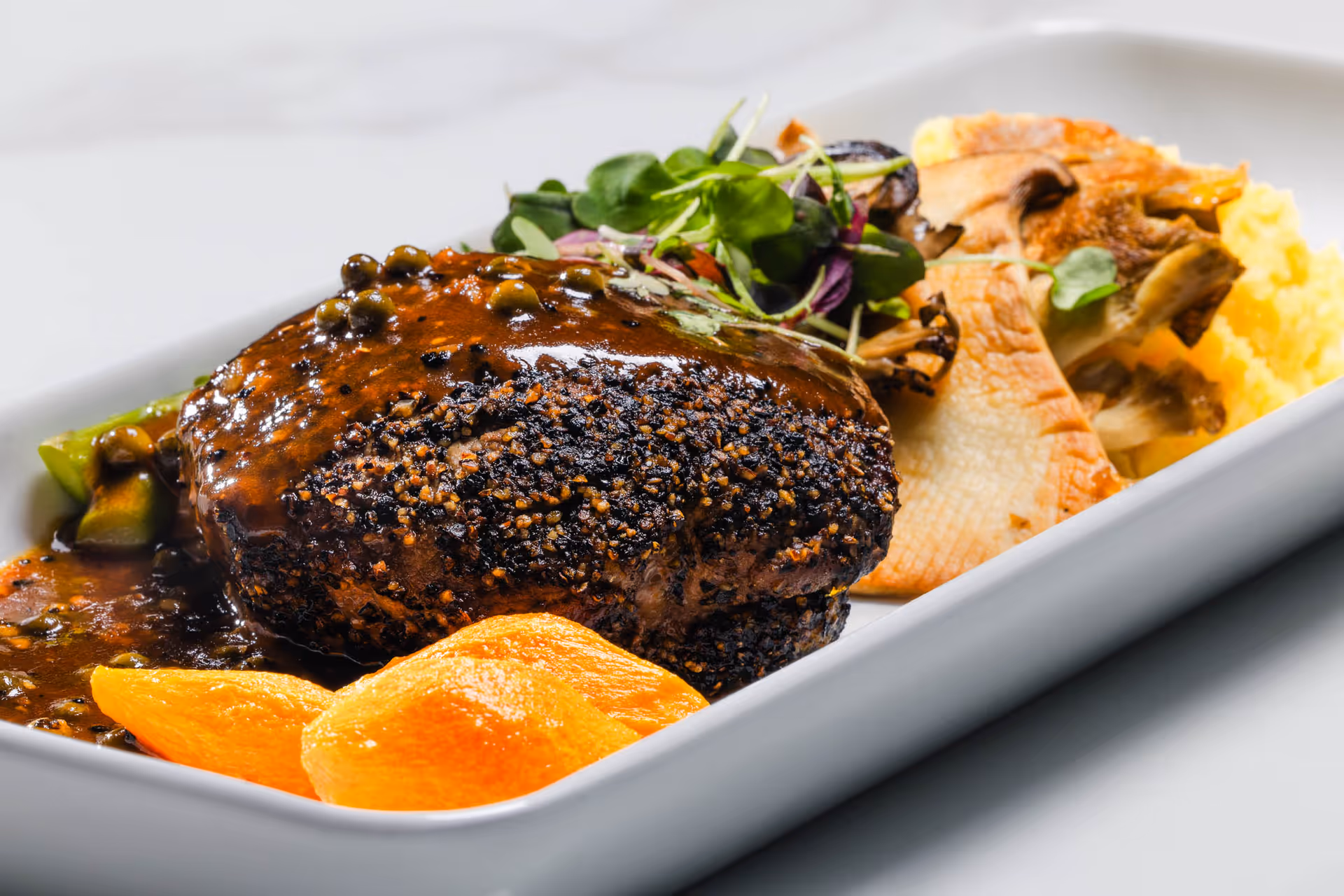 A gourmet dish featuring a pepper-crusted steak topped with a rich brown sauce and green peppercorns, served with roasted vegetables including mushrooms and orange-colored root vegetables, garnished with fresh greens on a white rectangular plate.