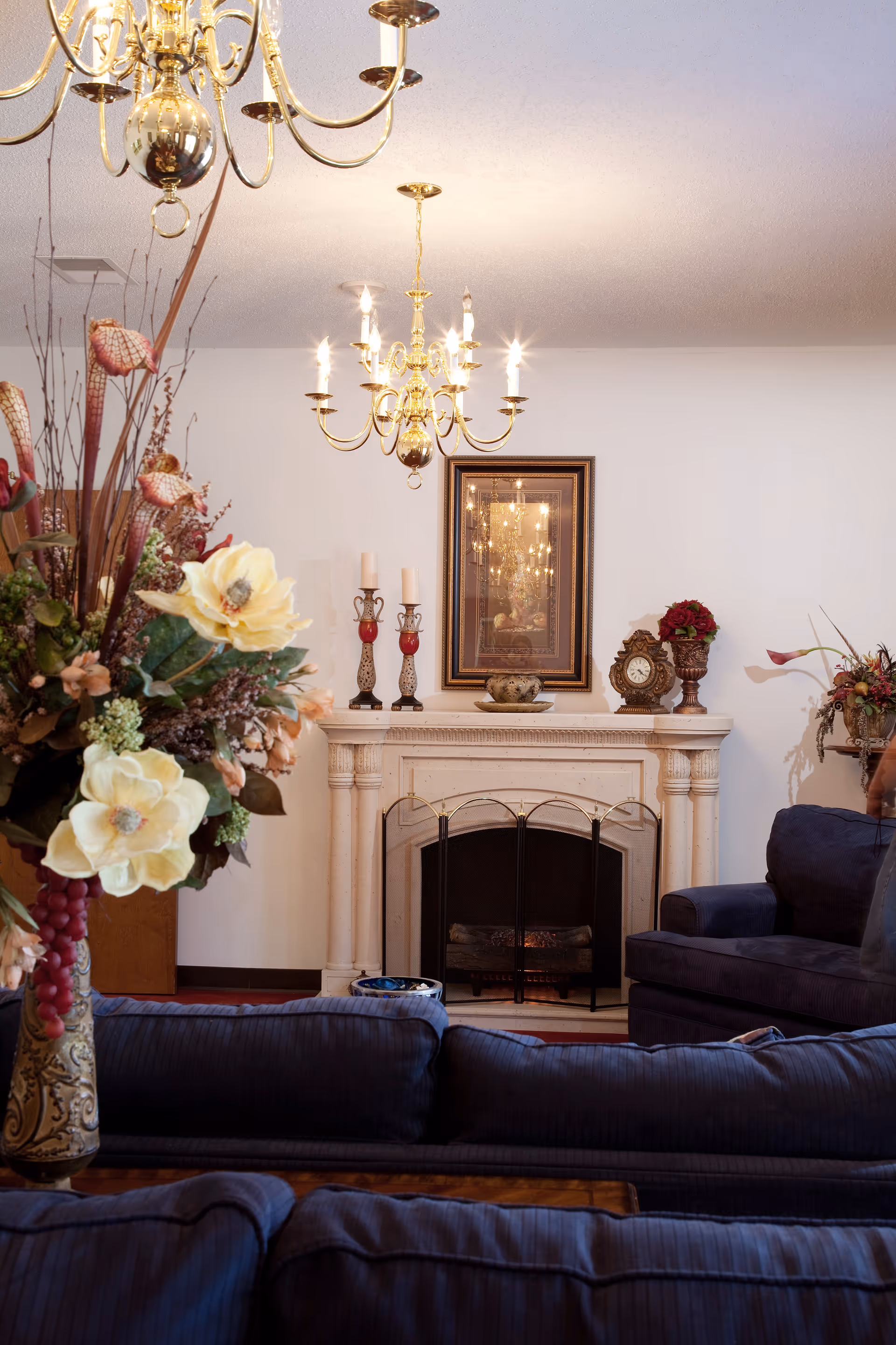 A cozy living room with a white fireplace adorned with candles, a clock, and floral decorations. Above the fireplace hangs a framed mirror reflecting a chandelier with lit bulbs. In the foreground, there are dark blue cushioned sofas and a large floral arrangement in a decorative vase.
