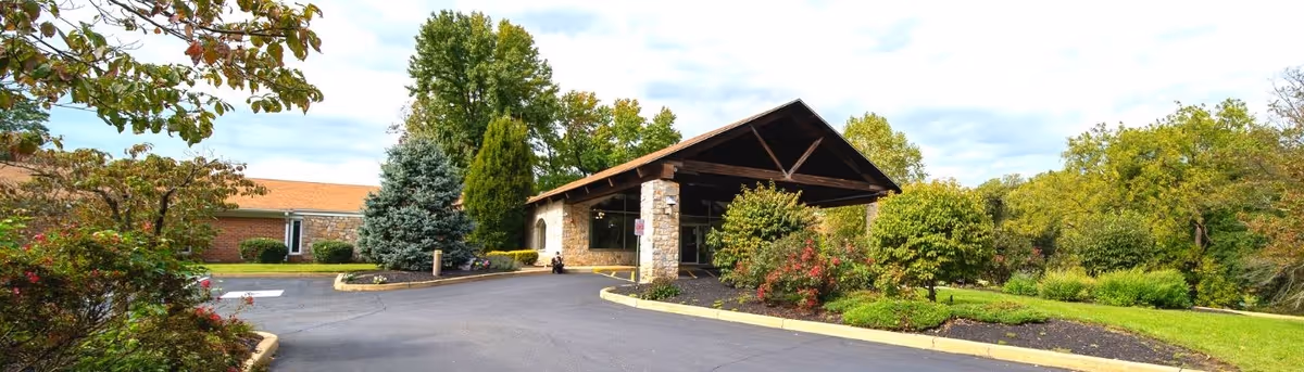 Exterior view of Symphony Manor of Feasterville showing a driveway leading to a covered entrance with stone pillars, surrounded by green trees and landscaped bushes under a partly cloudy sky.