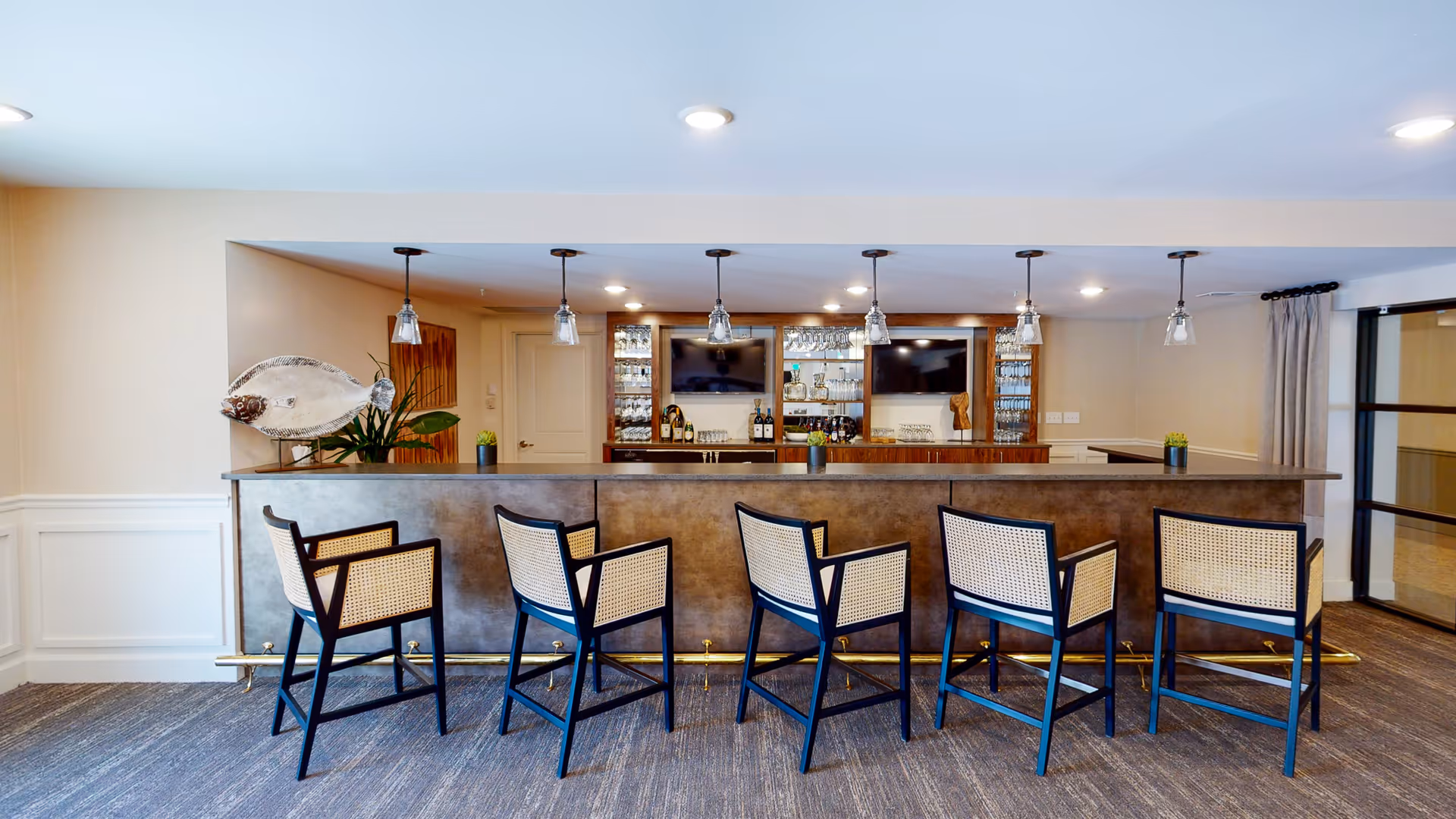 A modern indoor bar area with five cane-backed bar stools lined along a long counter, pendant lights overhead, and a mirrored back bar with bottles and glassware.