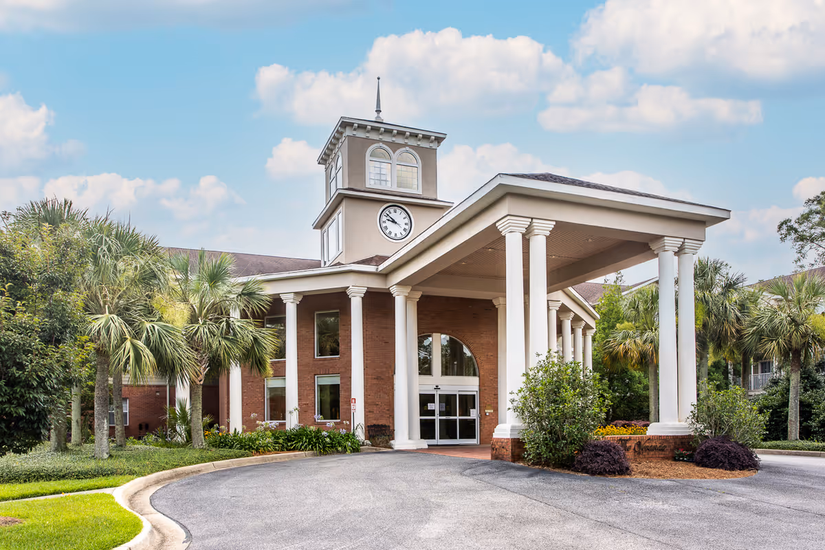 Exterior view of a senior living facility building with a covered entrance supported by white columns, a clock tower above the entrance, surrounded by palm trees and greenery under a partly cloudy sky.