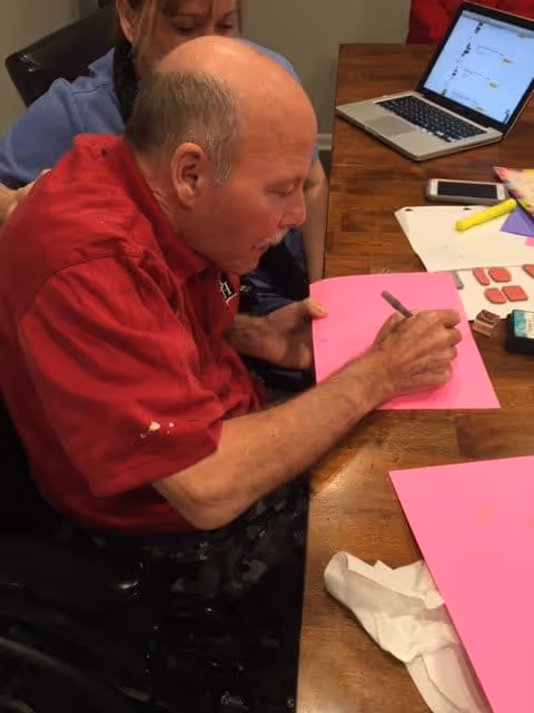 An elderly man in a red shirt is seated at a wooden table, writing or drawing on a pink sheet of paper with a pen. A woman is partially visible behind him, assisting or observing. On the table are additional pink sheets, a smartphone, a laptop, and some stationery items including highlighters and erasers.
