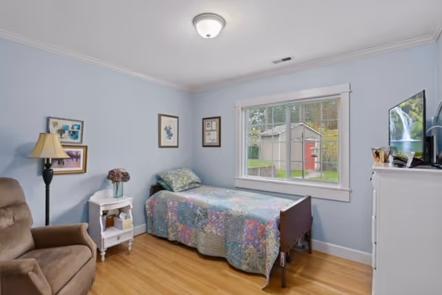 A small bedroom with light blue walls and wooden floor. The room contains a single bed with a colorful patchwork quilt, a brown armchair, a white nightstand with a lamp and a vase of flowers, and a white dresser with a TV on top. There are framed pictures on the walls and a window showing a view of a backyard with a shed.