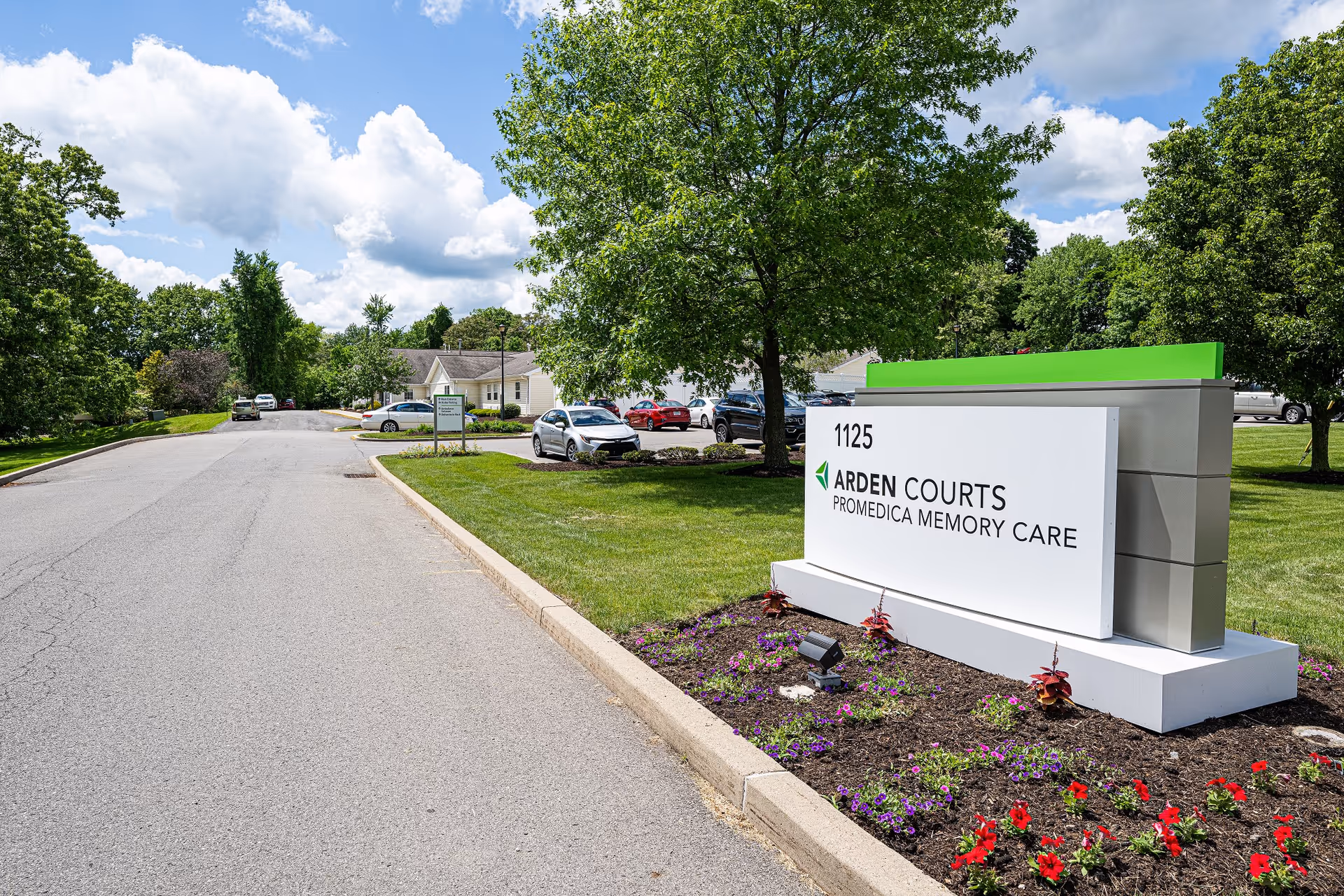 Outdoor view of Arden Courts - ProMedica Memory Care Community sign with the address 1125, surrounded by a landscaped flower bed. In the background, there is a parking area with several cars and trees under a partly cloudy blue sky.