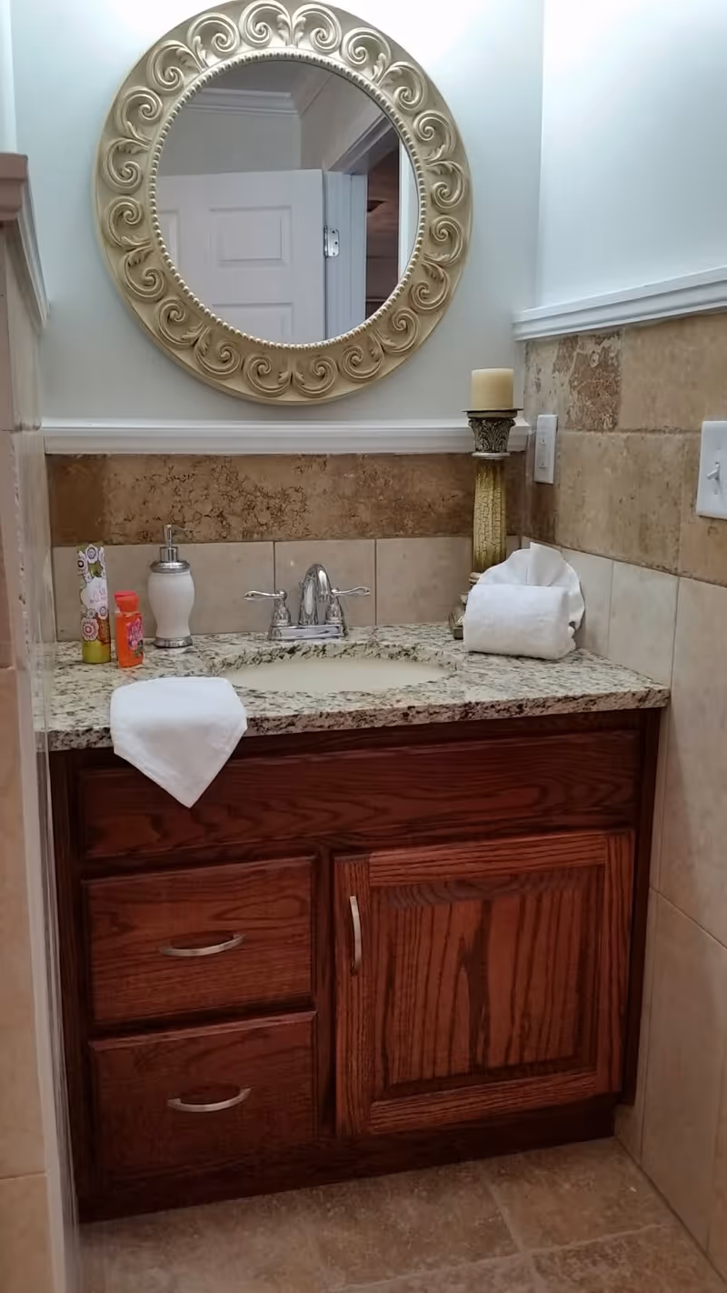 A bathroom vanity with a granite countertop, a silver faucet, and a decorative round mirror with an ornate frame above it. On the countertop, there is a soap dispenser, a candle on a tall holder, a rolled white towel, and some small toiletry items. The vanity has dark wood cabinets and drawers, and the walls are tiled halfway up with beige tiles.