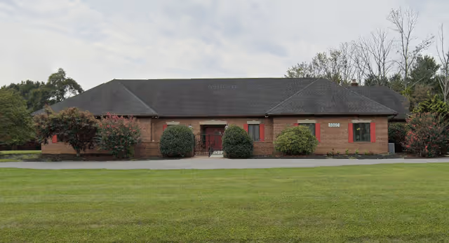 Single-story brick building with a dark shingled roof, red window shutters, and bushes in front, set against a cloudy sky and a large grassy lawn.