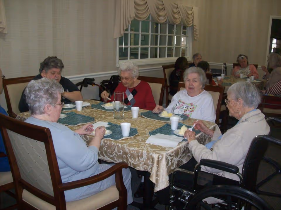 A group of elderly women sitting around a table in a dining room, eating and conversing. The table is covered with a patterned tablecloth and set with placemats, plates of food, and white disposable cups. The room has beige walls, a window with draped curtains, and wooden chairs. One woman is in a wheelchair.