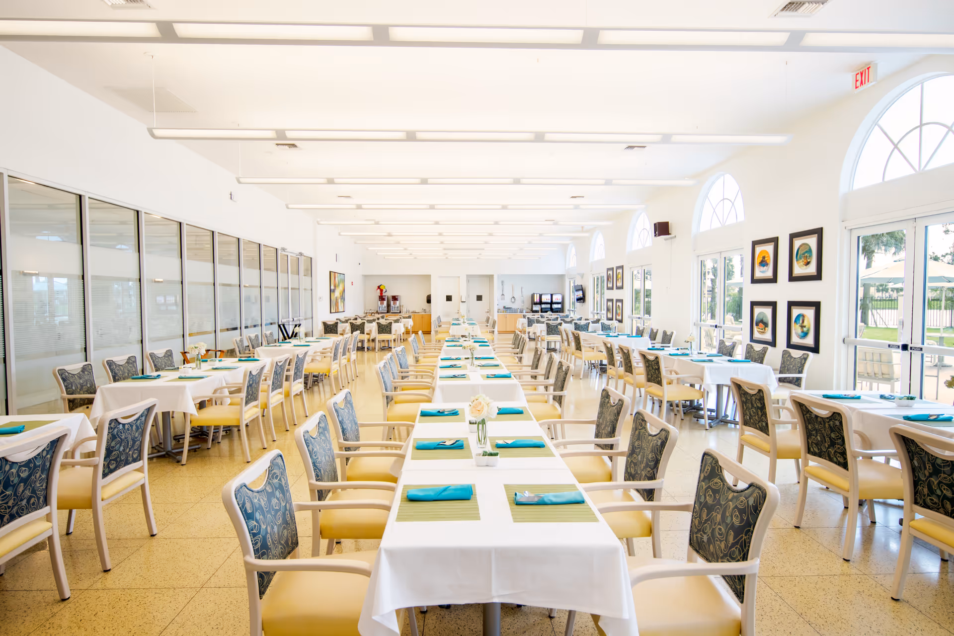 A bright and spacious dining room with multiple tables covered in white tablecloths, each set with green placemats and turquoise napkins. The room features large arched windows letting in natural light, framed artwork on the walls, and comfortable chairs with patterned backs and yellow seats arranged neatly around the tables.