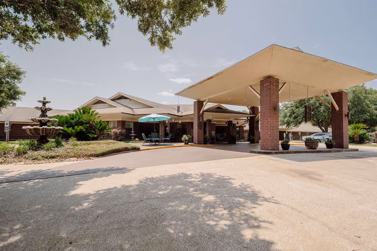 Exterior view of Silsbee Oaks Health Care facility showing the entrance with a covered drop-off area supported by brick columns, a multi-tiered water fountain on the left, outdoor seating with a green umbrella, and surrounding greenery under a clear sky.