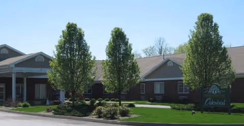 Exterior view of Oakwood Health Campus showing a single-story brick building with a covered entrance, three tall trees, and a green lawn with landscaping in front. A sign with the facility name is visible on the right side of the image.