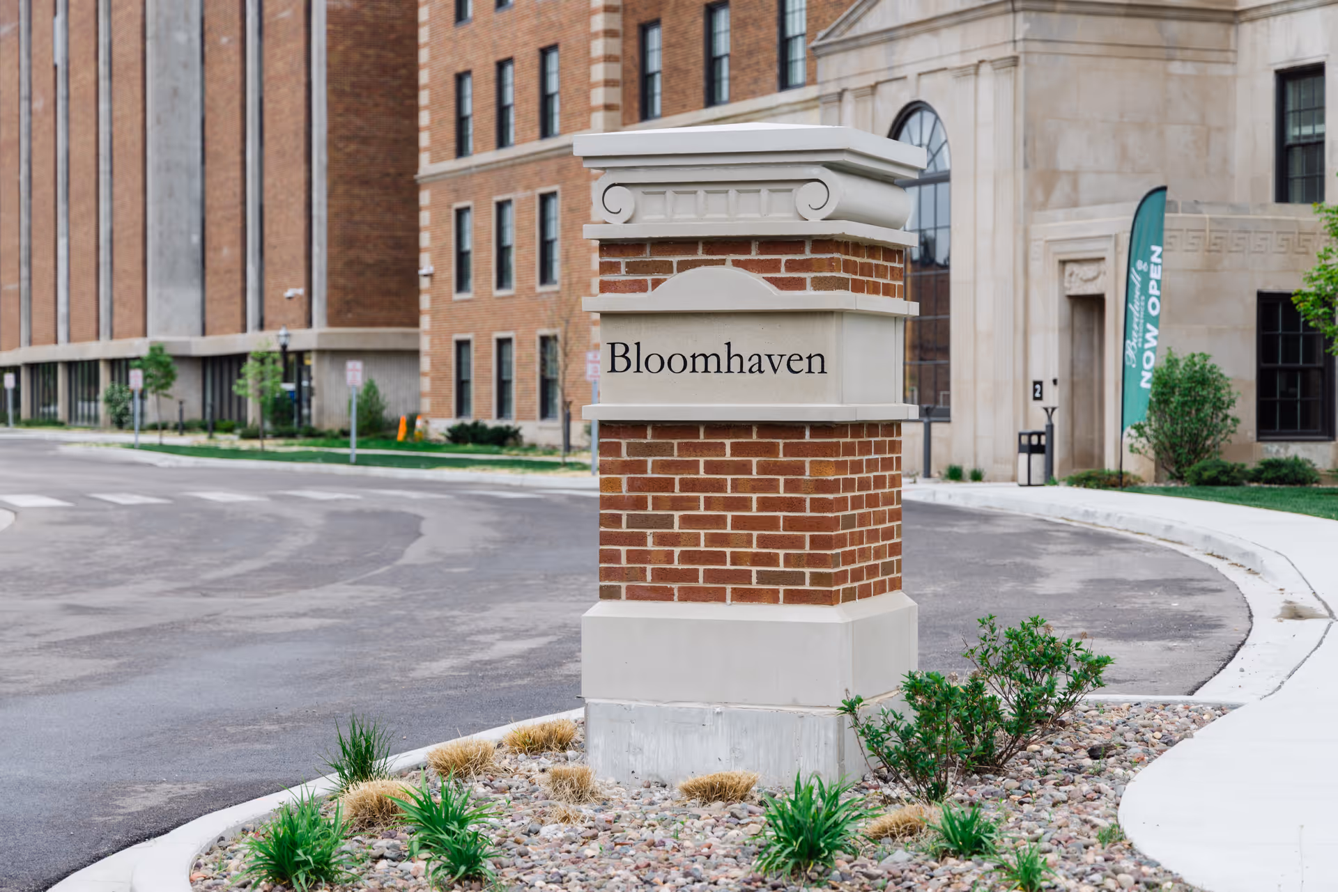 A brick and stone monument sign with the name 'Bloomhaven' on it, situated at the entrance of a residential building complex. The background shows a multi-story brick building and a green flag with the text 'NOW OPEN'. There are small plants and rocks around the base of the sign.