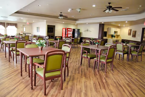 A spacious dining room with multiple wooden tables and green cushioned chairs arranged neatly. The room has wood flooring, ceiling fans, vending machines against the far wall, and soft lighting. There are decorative plants on some tables and framed artwork on the walls.
