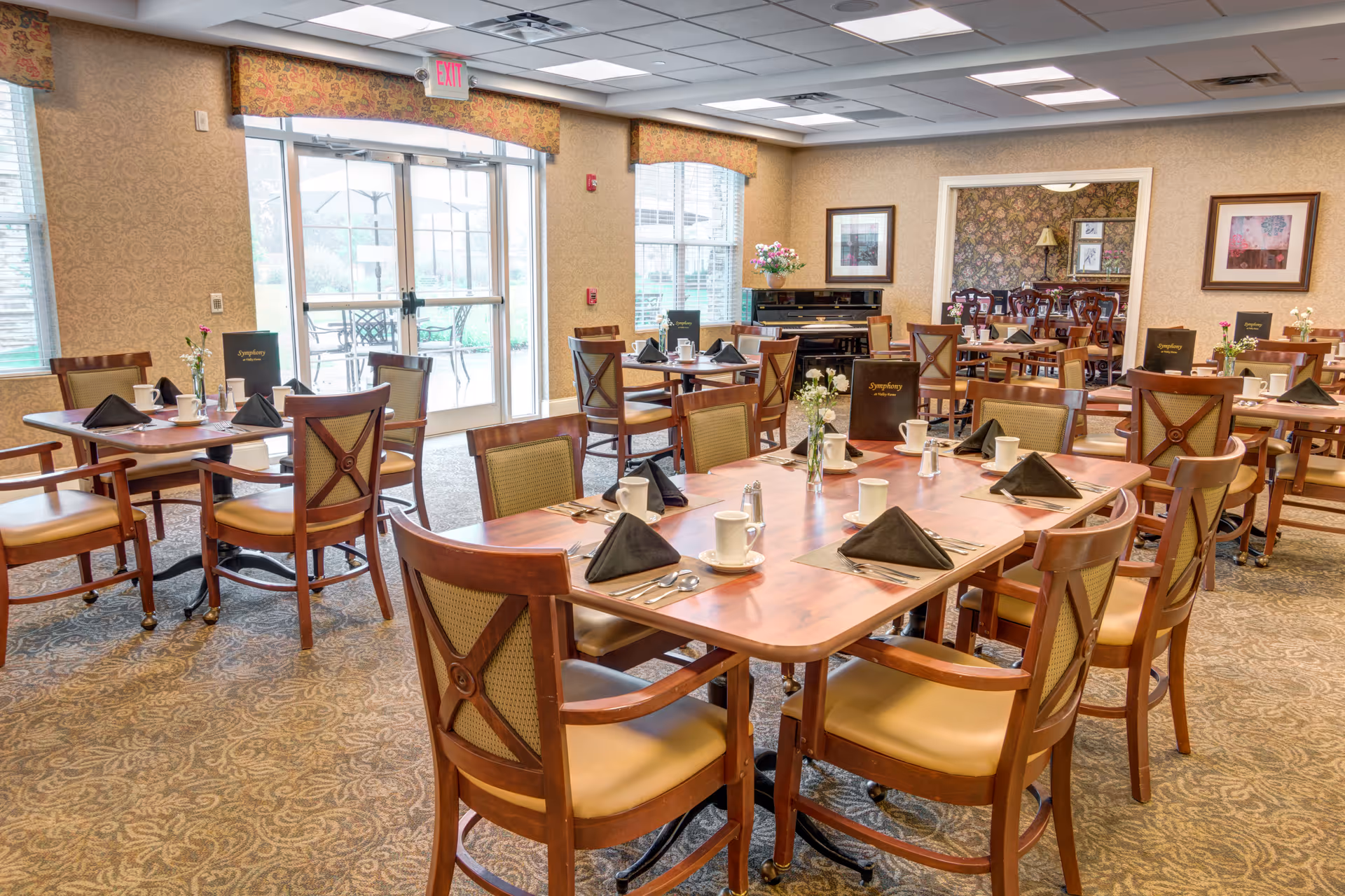 A bright and spacious dining room with multiple wooden tables and chairs arranged neatly. Each table is set with white cups, black napkins, silverware, and small flower vases. Large windows and glass doors allow natural light to fill the room, and framed artwork decorates the walls. The room has a warm and inviting atmosphere.