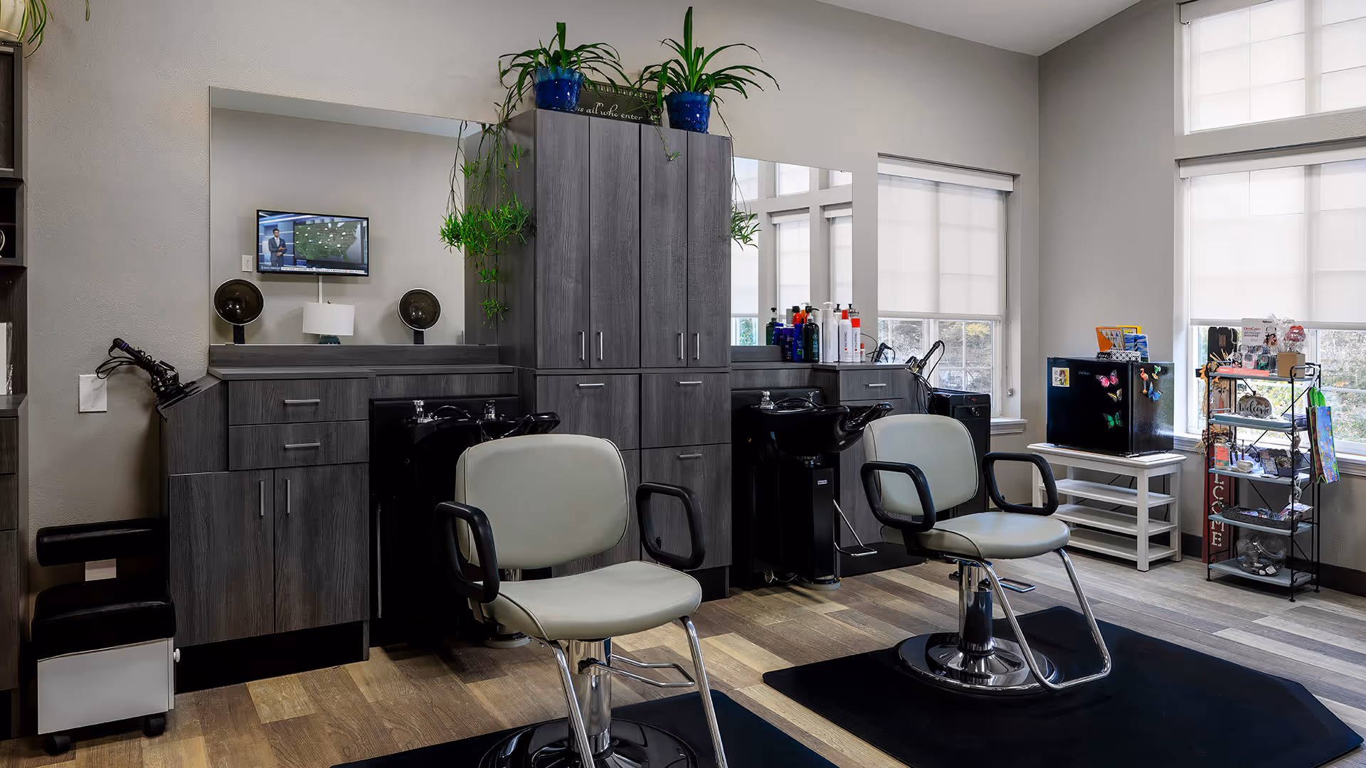 Interior view of a salon area in a senior living facility with two salon chairs in front of black hair washing sinks. There are gray cabinets with plants on top, a large mirror, and a small TV mounted on the wall. The room has large windows with white blinds, a small black refrigerator, and a metal rack with various items on it.