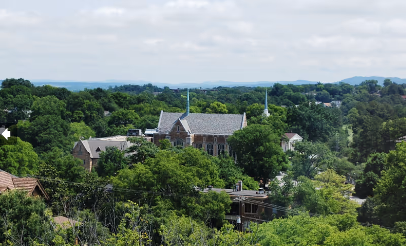 Aerial view of a building surrounded by dense green trees with hills visible in the background under a partly cloudy sky.