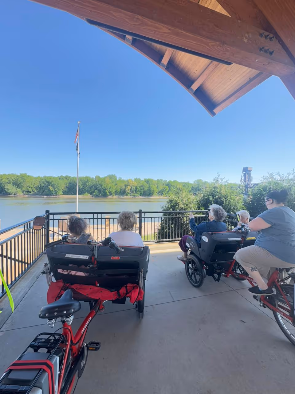 Four elderly individuals sitting on two red tandem bikes with seats designed for passengers, under a wooden roof structure, overlooking a river with trees on the opposite bank and a flagpole with an American flag.