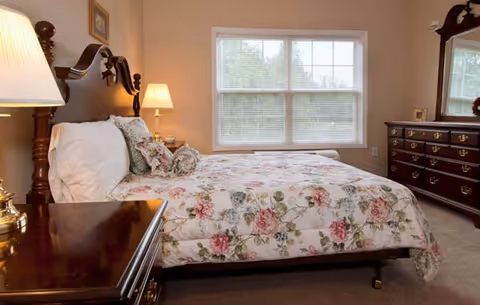 Sunlit bedroom with a floral bedspread, wooden headboard, nightstands with lamps, dresser, and a window.