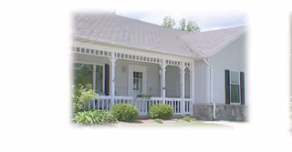 Exterior view of a single-story white residential building with a covered front porch, decorative trim, and a shingled roof. There are shrubs and greenery in front of the porch.