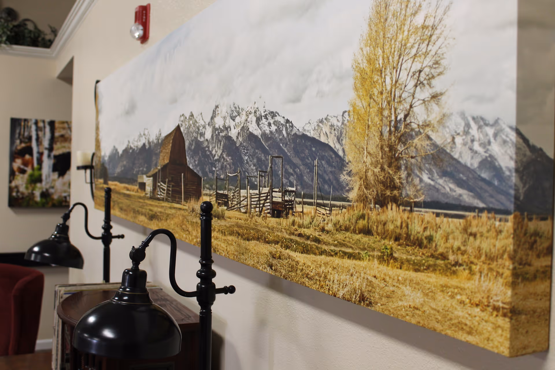 Large landscape canvas print of a barn and mountains mounted on an interior wall above black reading lamps and seating.