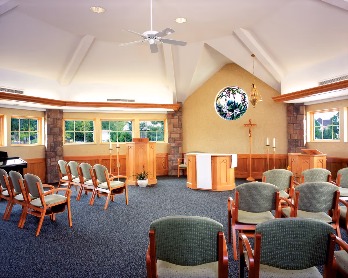 Interior of a chapel or small worship room with rows of green cushioned chairs, wooden podiums, a cross, and a round stained glass window depicting flowers. The room has a high vaulted ceiling with a ceiling fan and large windows letting in natural light.