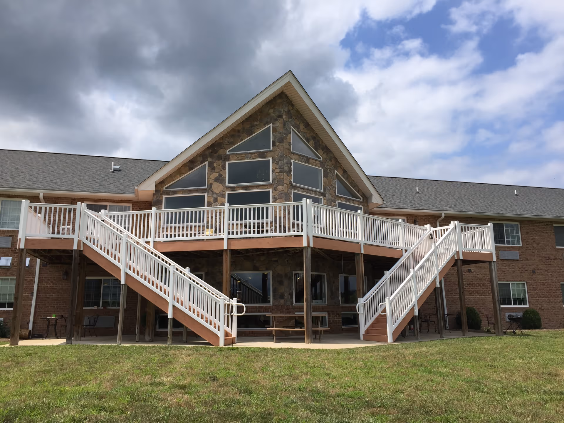 Exterior view of a two-story senior living facility building with a large stone facade section featuring multiple triangular and rectangular windows. There is a wooden deck with white railings and two staircases leading down to a grassy lawn. The sky is partly cloudy.