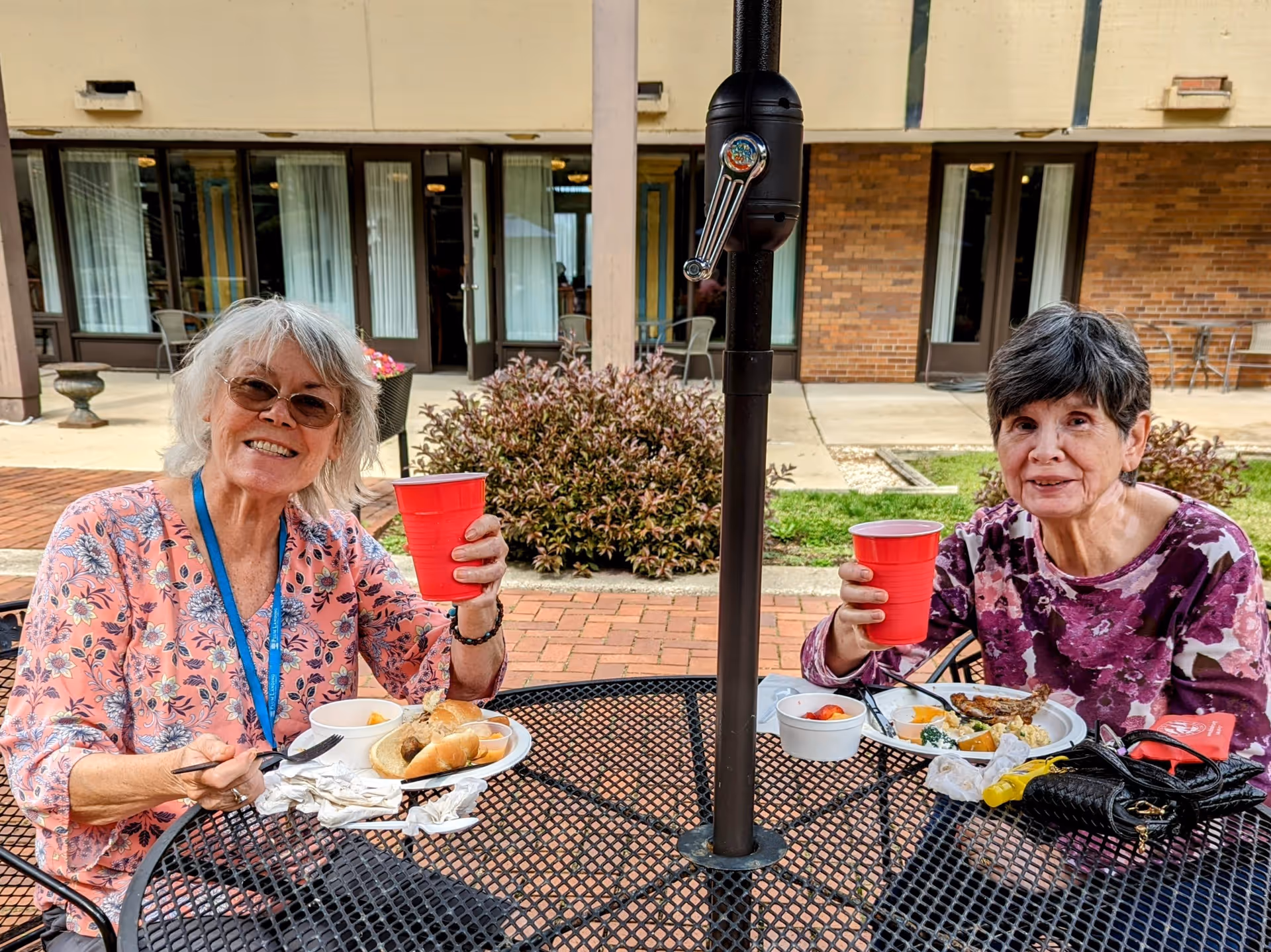 Two elderly women sitting at a black metal outdoor table enjoying a meal and holding red plastic cups. They are smiling and appear to be in a courtyard area with brick paving, plants, and building doors in the background.