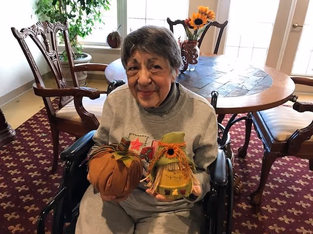 An elderly woman sitting in a wheelchair inside a room with a round wooden table and chairs. She is holding two small decorative fall-themed items, one resembling a pumpkin and the other a scarecrow. There is a potted plant and a vase with sunflowers on the table in the background.