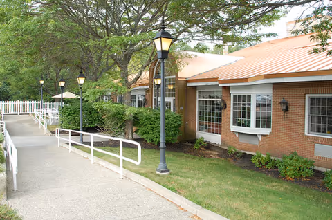 Exterior view of a brick building with large windows and a sloped roof, surrounded by green bushes and trees. A paved walkway with white railings and several lamp posts leads up to the entrance.