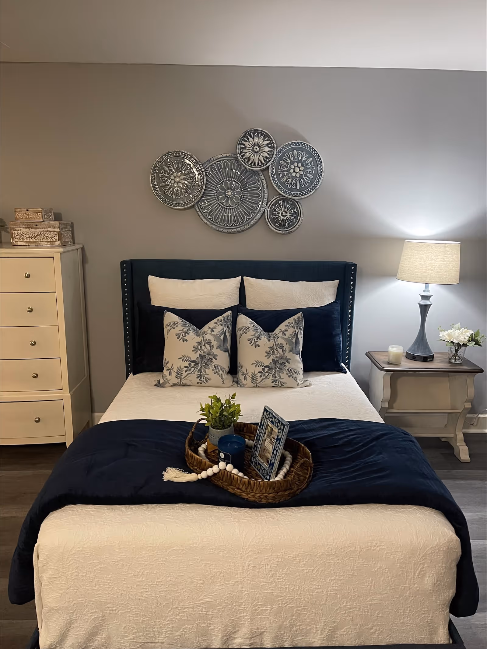 A neatly made bed with white and navy blue bedding, decorated with floral and solid pillows. Above the bed, there is a wall decoration consisting of five round, ornate metal plates. To the left of the bed is a cream-colored dresser with three drawers and decorative boxes on top. To the right is a small nightstand with a lamp, a candle, and a small flower arrangement. The room has gray walls and wood flooring.