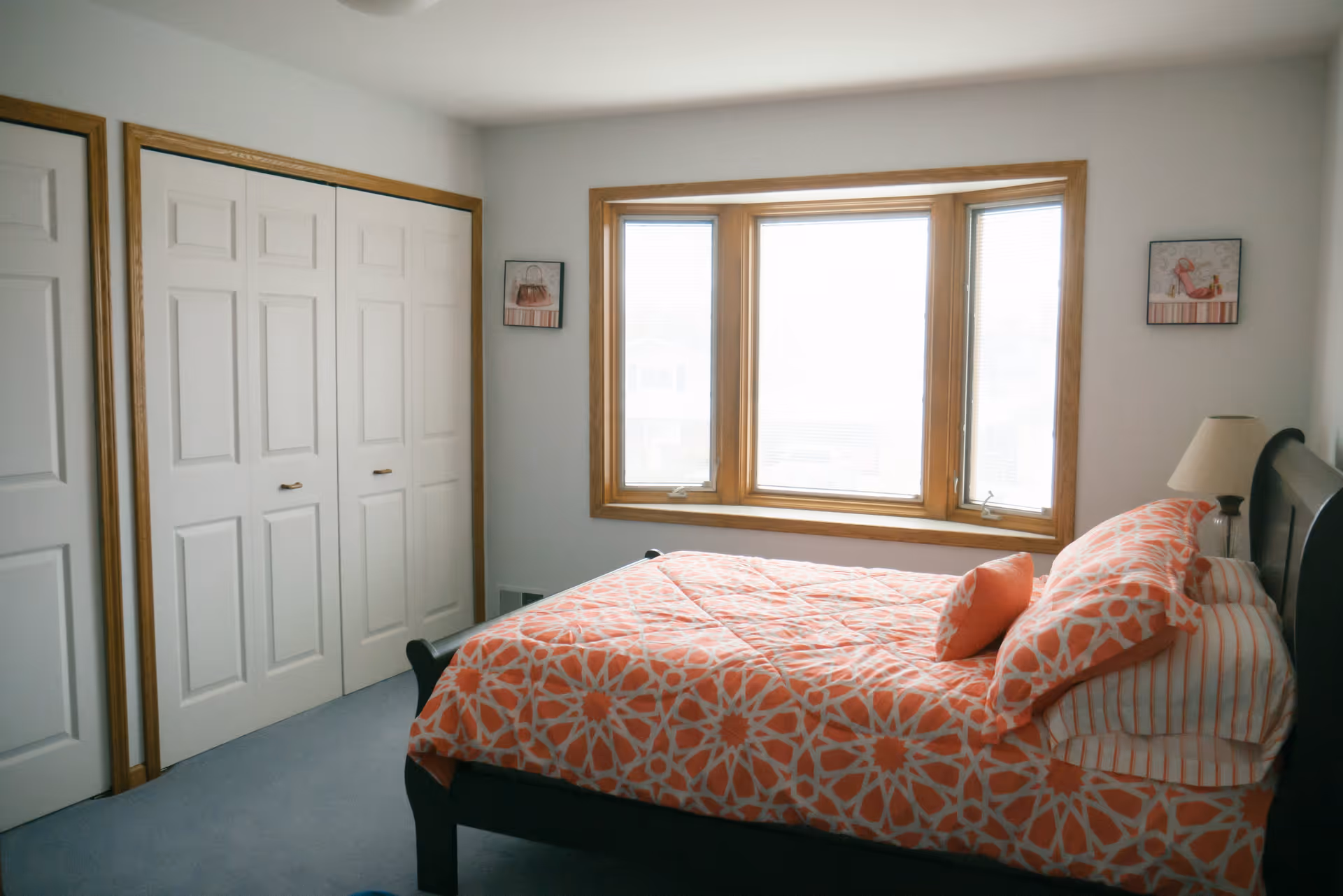 A bedroom with a bed covered in orange and white patterned bedding, a wooden framed window with blinds, two small framed pictures on the wall, a bedside table with a lamp, and white closet doors with wooden trim.