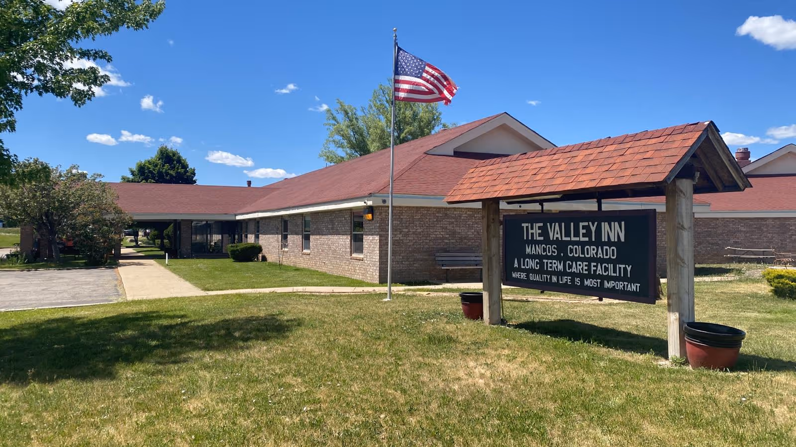 Exterior view of The Valley Inn, a long term care facility in Mancos, Colorado, showing a single-story brick building with a red roof, an American flag on a flagpole, a covered sign with the facility's name, and a grassy lawn under a clear blue sky.