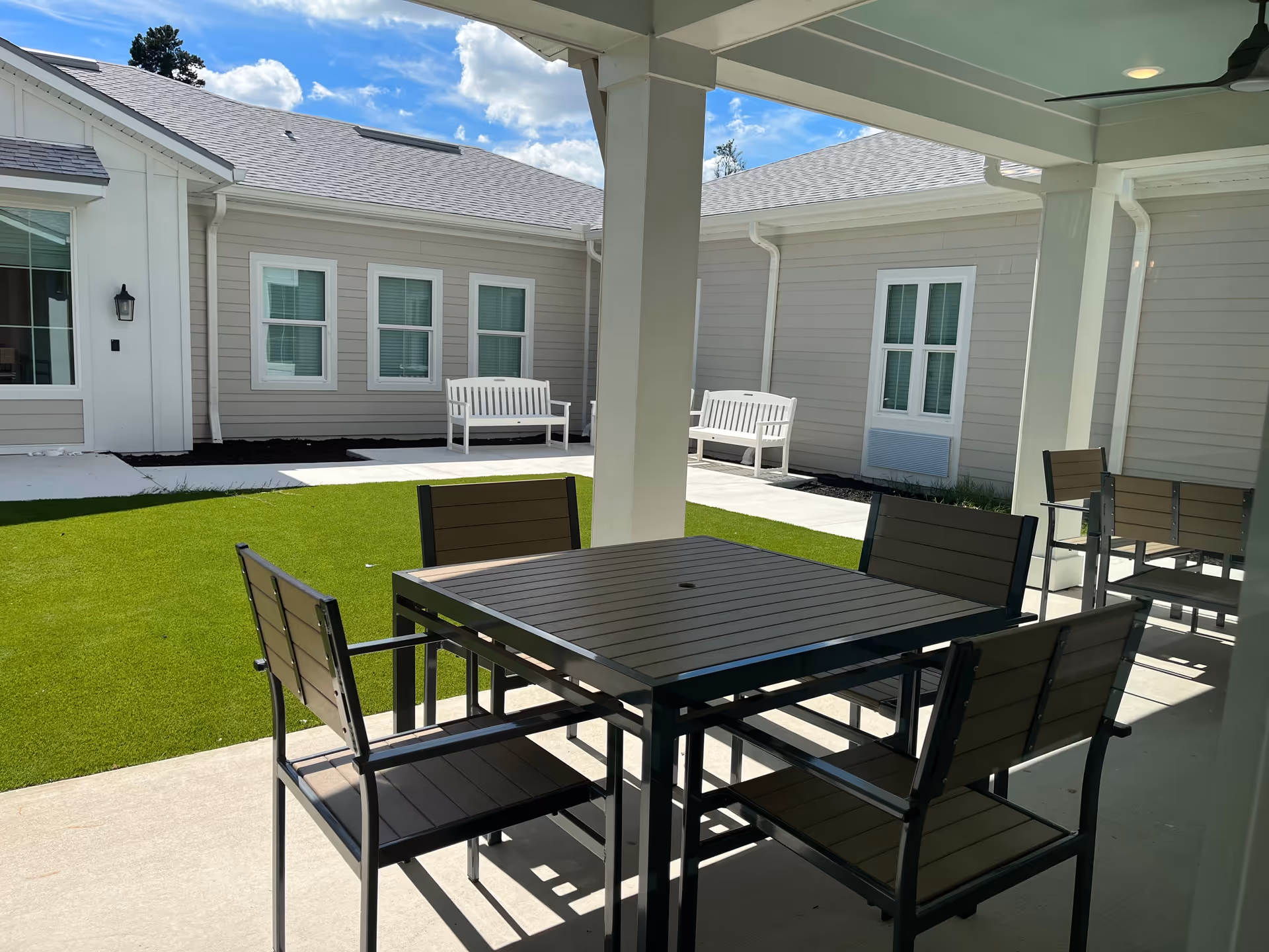 Outdoor covered patio area with a square table and four chairs, overlooking a courtyard with green artificial grass, concrete walkways, and white benches against the beige exterior walls of the building under a partly cloudy sky.